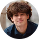 Young man with curly brown hair and a slight smile wearing a dark jacket against a rustic background.
